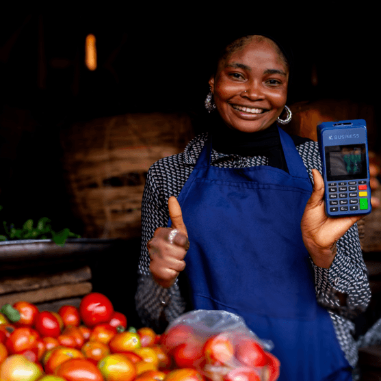 Smiling market vendor with fresh tomatoes, holding a handheld POS terminal and giving a thumbs up.